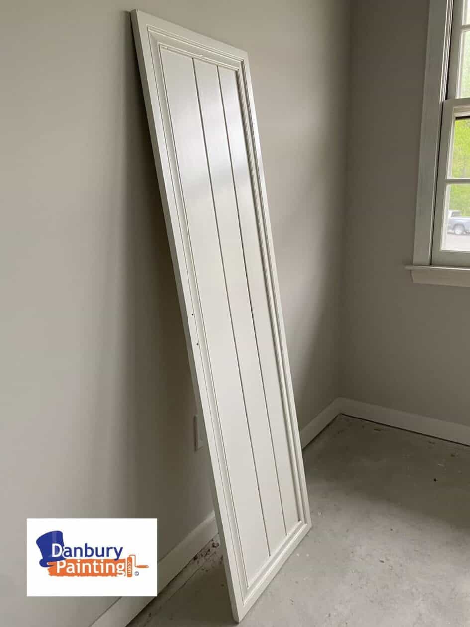White interior panel door leaning against a beige wall in an unfinished room near a window; Danbury Painting logo visible in the corner.